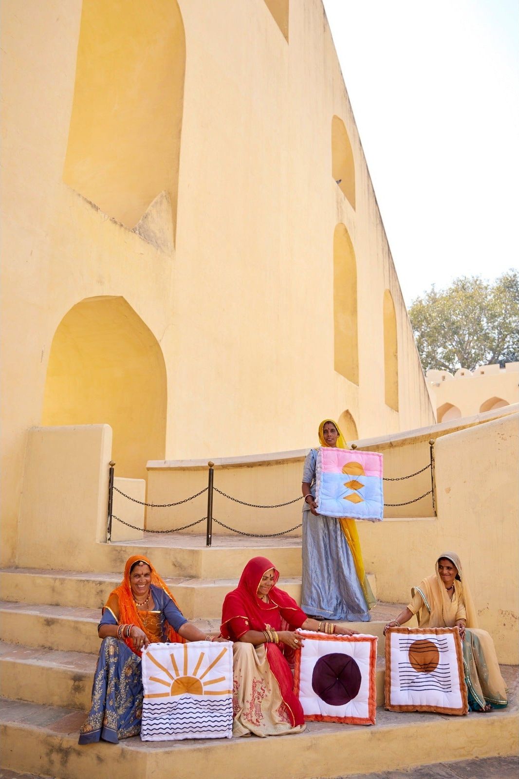 Three women sitting on steps in front of a large beige building, each holding a colorful fabric floor cushion that they handmade