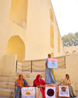 Three women sitting on steps in front of a large beige building, each holding a colorful fabric floor cushion that they handmade