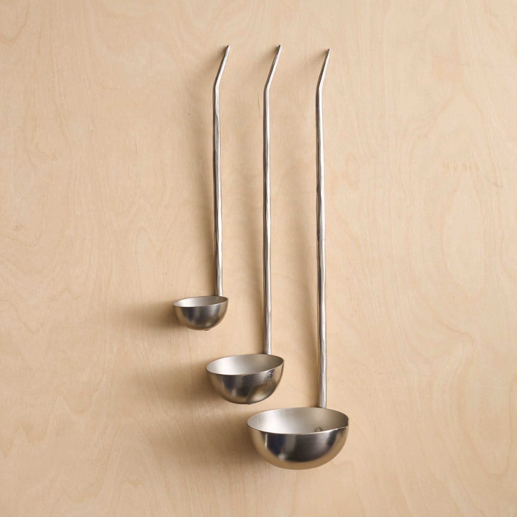 A set of three assorted sizes of pewter ladles with roomy cups and hammered handles, displayed against a wooden background