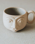 A beige ceramic mug with a face design on the front
