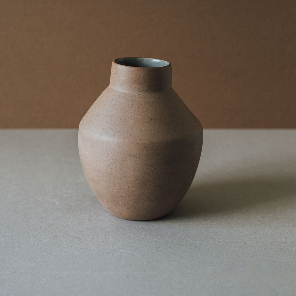 A handcrafted ceramic vase with a rounded body and a narrower top, presented in a muted brown color, placed on a flat surface.