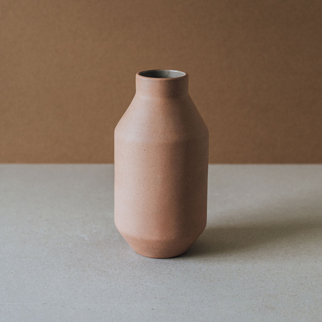 A cylindrical brown ceramic vase with a narrower top and wider base, placed on a flat surface with a plain background.