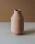A cylindrical brown ceramic vase with a narrower top and wider base, placed on a flat surface with a plain background.