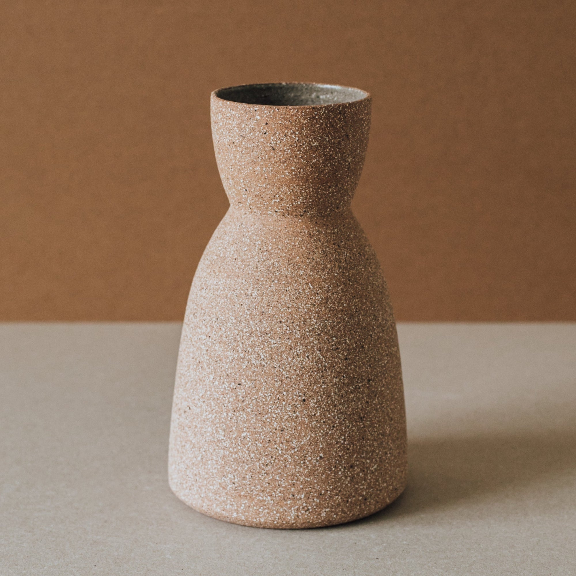 A beige handcrafted ceramic vase with an hourglass shape, made from a mixture of local clay, displayed against a neutral background.