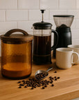 Coffee-making setup with a canister, French press, grinder, and mugs on a wooden surface.