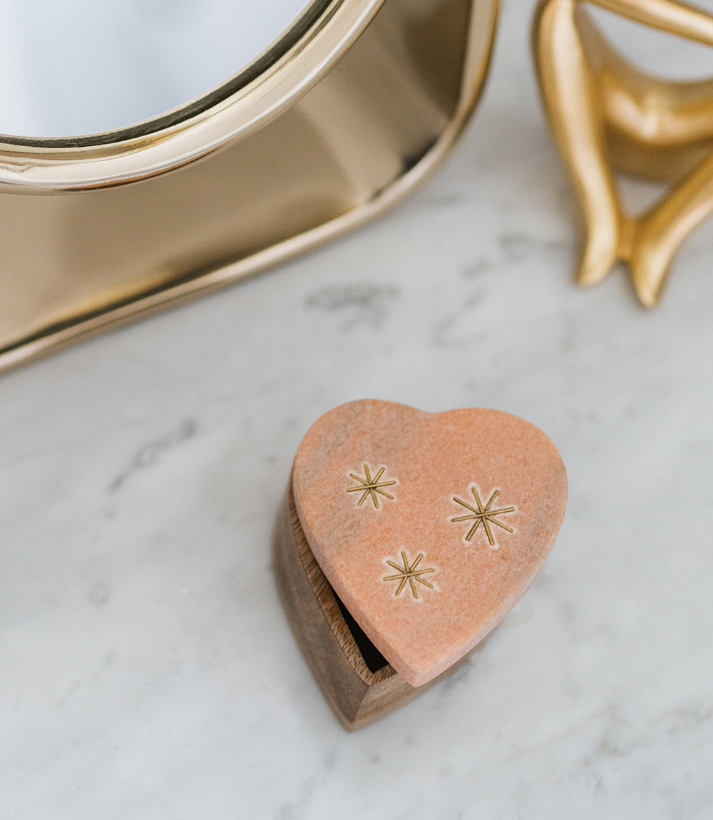 A heart-shaped pink marble keepsake box with brass stars inlay on the lid, set on a wooden base on a marble table