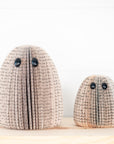 Two decorative book-folded ghost sculptures in different sizes, made from beige and brown book pages with black text, featuring black button eyes, displayed on a light wooden surface against a white paneled wall.