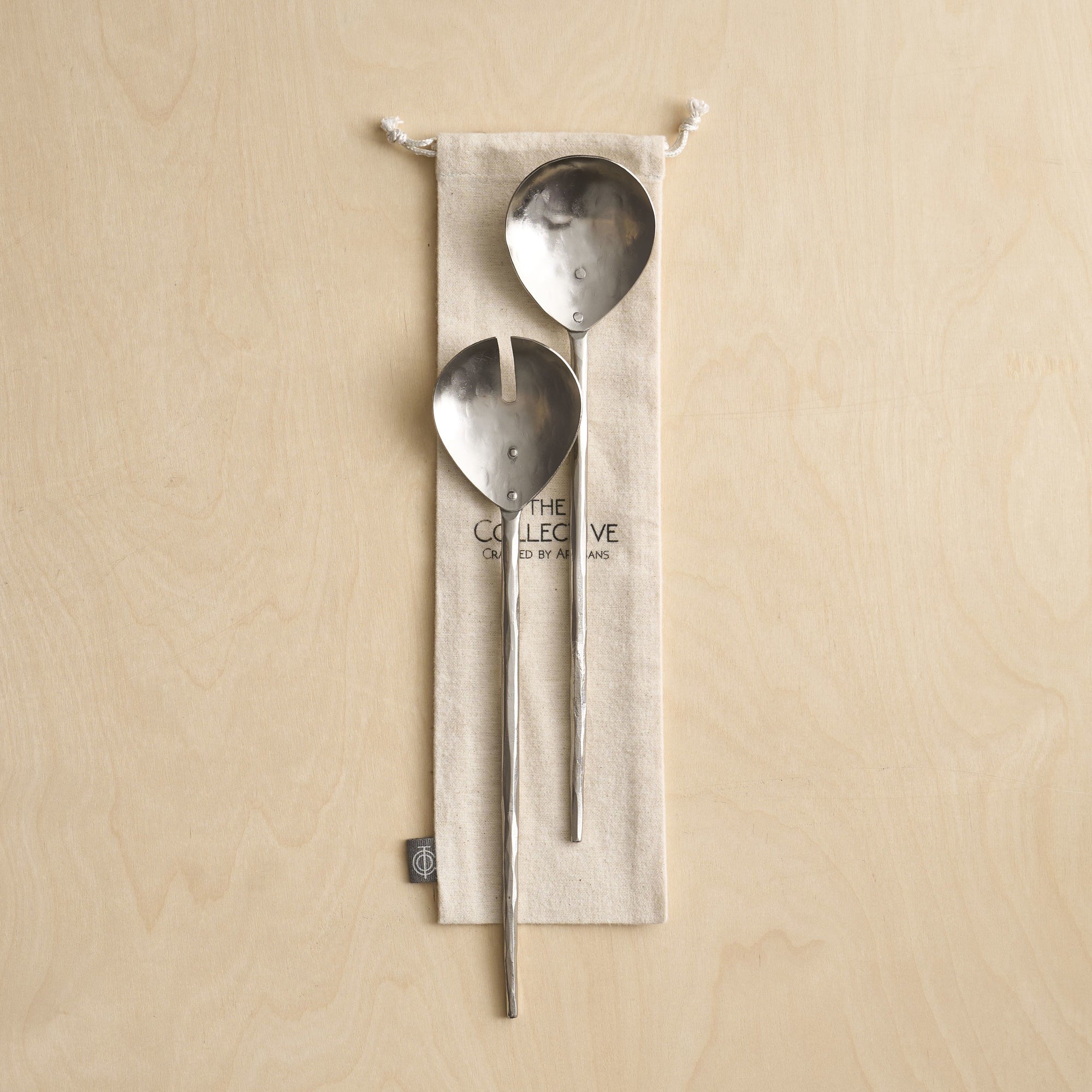 Two pewter salad servers with elongated, hammered handles, displayed on a beige linen cloth with the collective logo