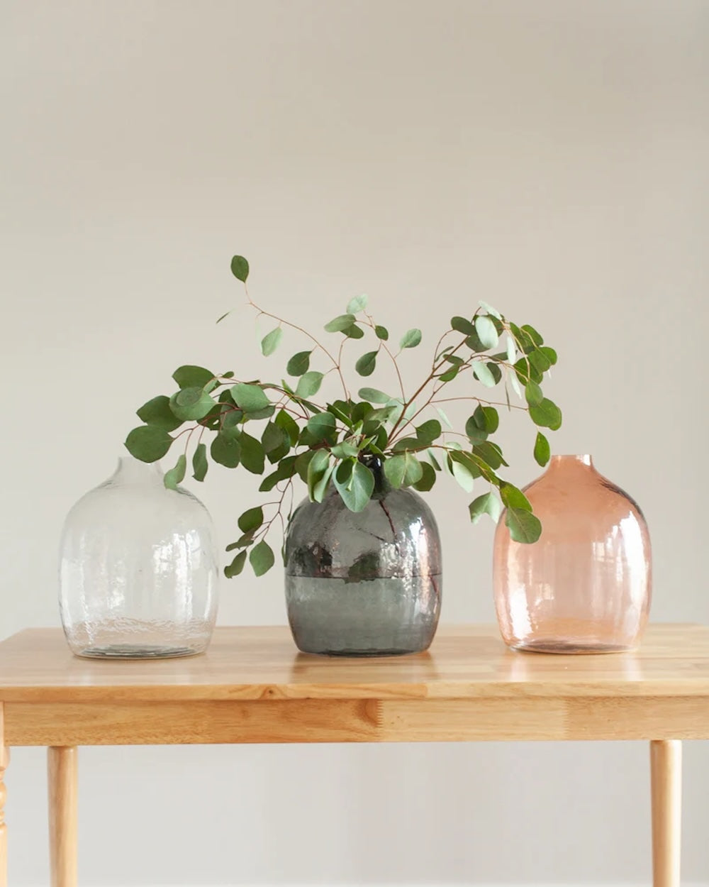 Three vases on a wooden table with greenery, one of which is transparent, another gray, and the third pink.