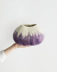 Hand holding a textured wool decorative bowl with a gradient from purple to beige against a white background