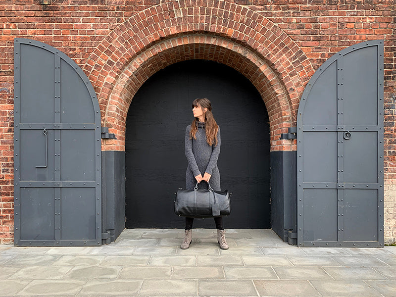 a female holding a black leather weekender with a leather handle and visible stitching details outside.