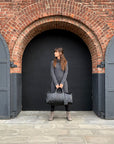 a female holding a black leather weekender with a leather handle and visible stitching details outside.