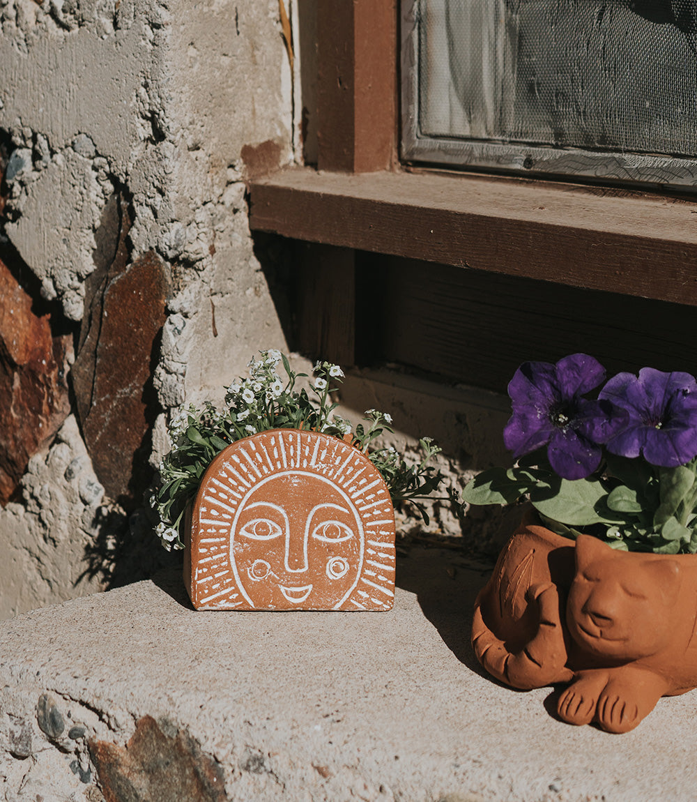 A handmade terracotta planter with a hand-painted sunrise design outside next to a cat planter