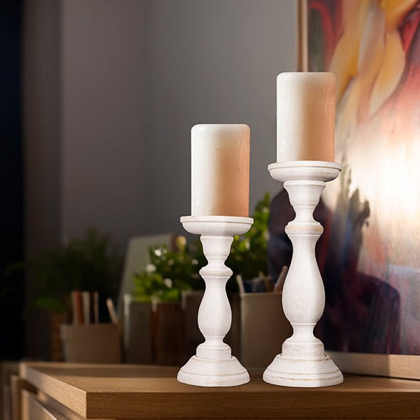 A set of two white shabby chic wooden candleholders in different sizes on a wooden table with a soy wax candle on top