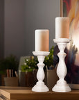 A set of two white shabby chic wooden candleholders in different sizes on a wooden table with a soy wax candle on top