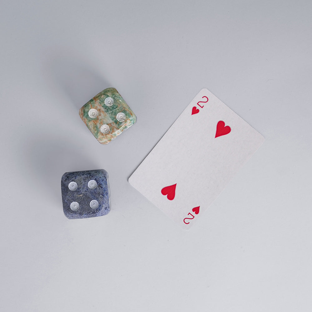 A pair of two hand-carved gemstone dice, in blue and multi-color, displayed on a white surface next to the two of hearts playing card.