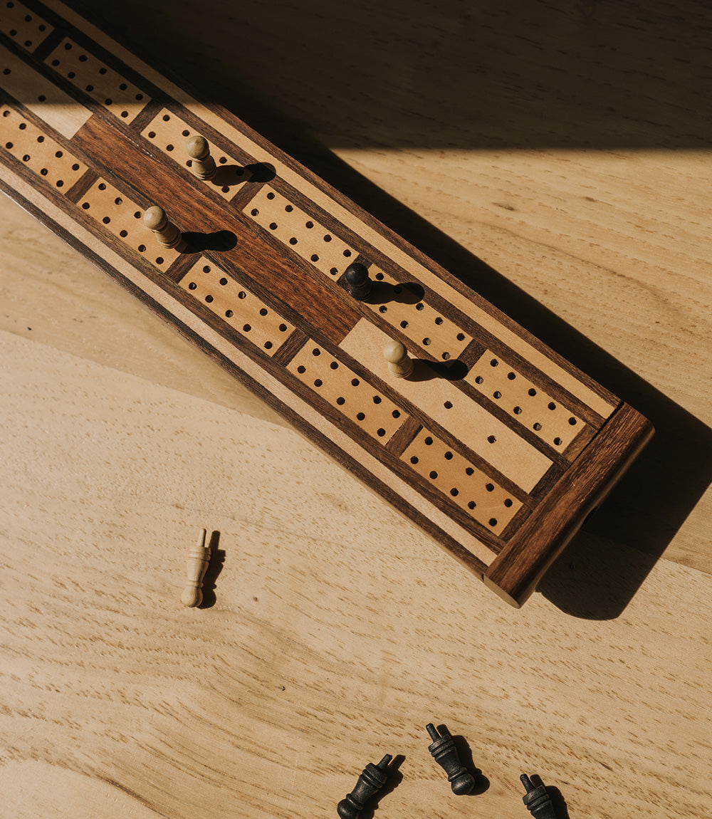 A traditional wooden cribbage board with carved-out storage for game pieces, featuring a patterned design with black and gold elements on a wooden table