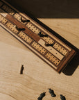 A traditional wooden cribbage board with carved-out storage for game pieces, featuring a patterned design with black and gold elements on a wooden table