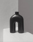 A black, handmade ceramic vase with an abstract shape, featuring a wide base that narrows at the neck, displayed against a neutral background.