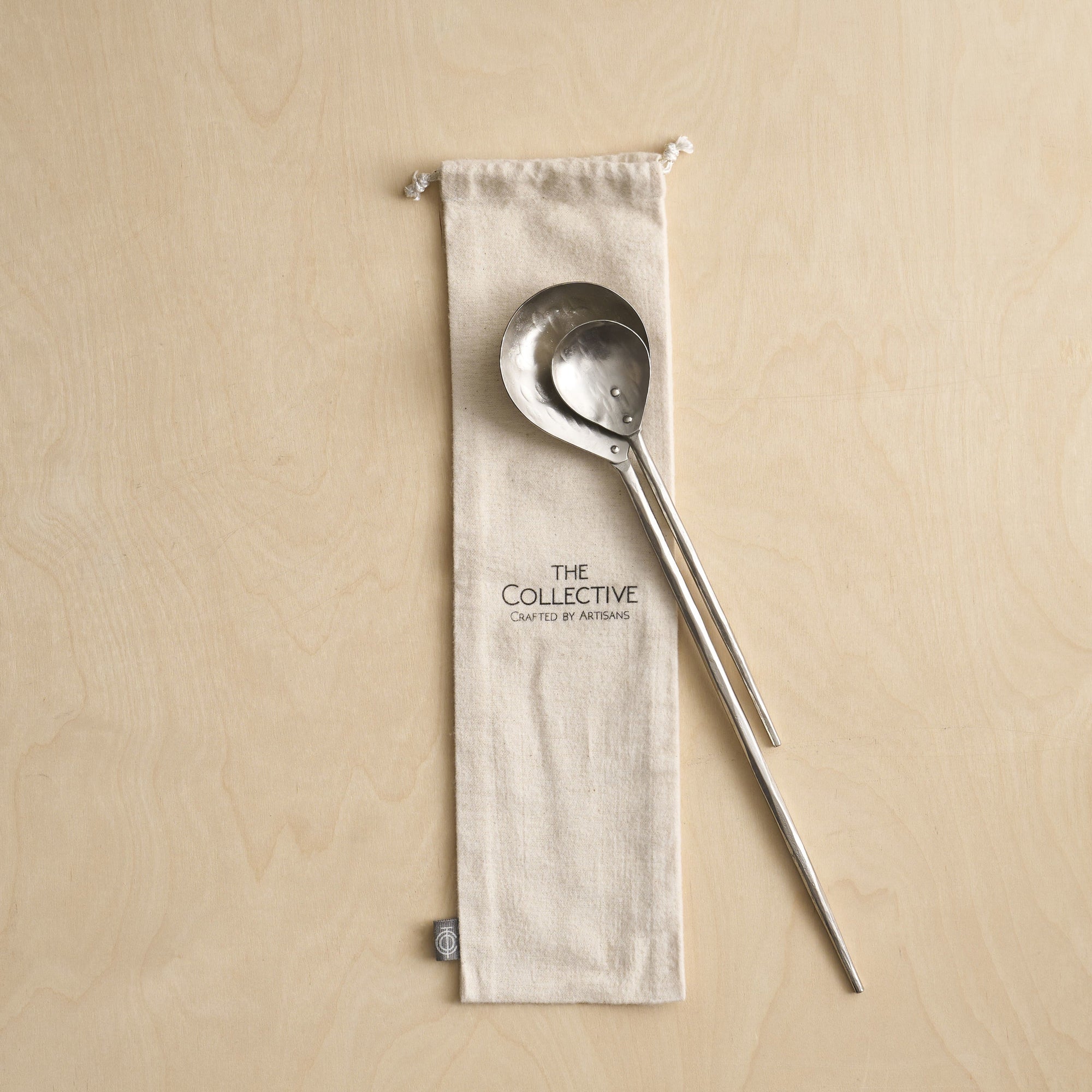 A set of two pewter stir spoons, one large and one small, with an elongated handle, placed on a natural color flannel bag with The Collective Home logo