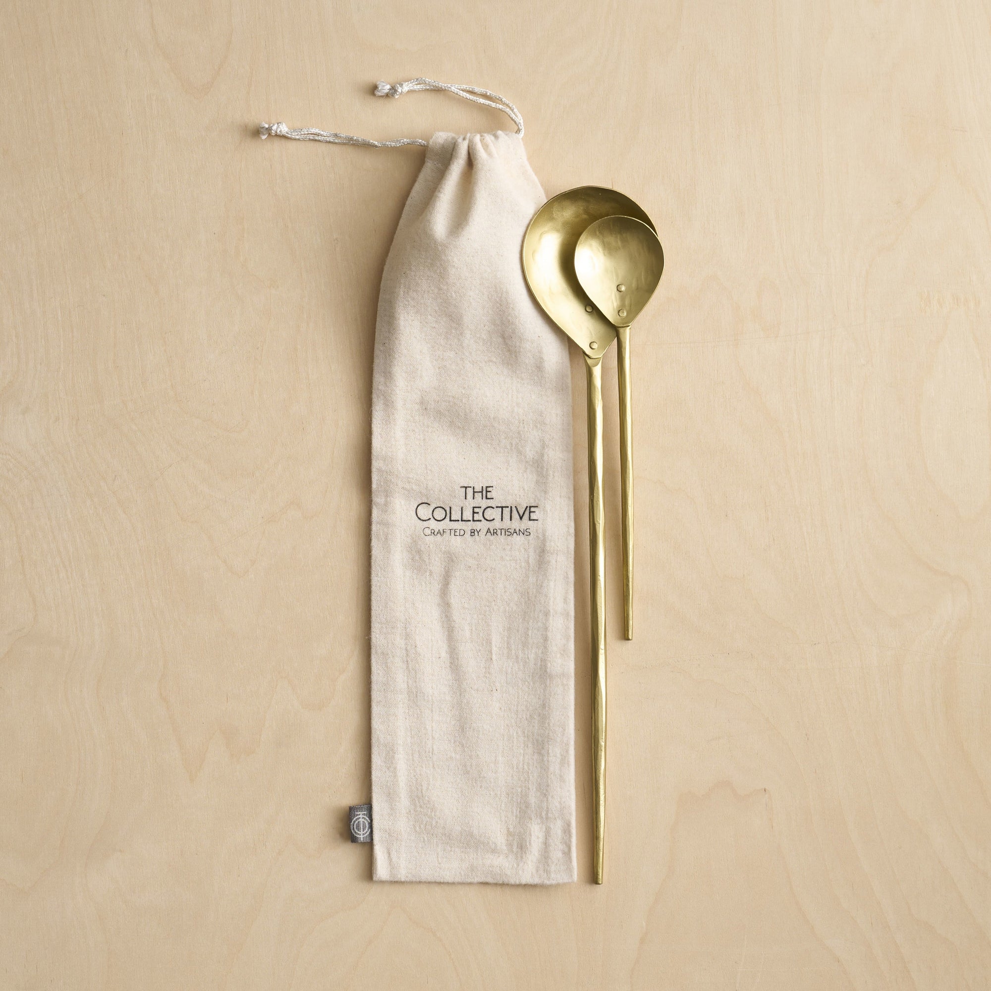 A set of two brass stir spoons with a matte finish, displayed alongside a beige cotton bag with 'The Collective' branding on it, placed on a wooden surface