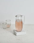 A clear glass pitcher with a hammered texture, filled with a pink liquid, placed on a white surface, with two empty glasses in the background
