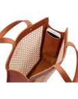 The inside of a brown leather tote bag with rattan cane webbing and leather handles with a phone in the side pocket.