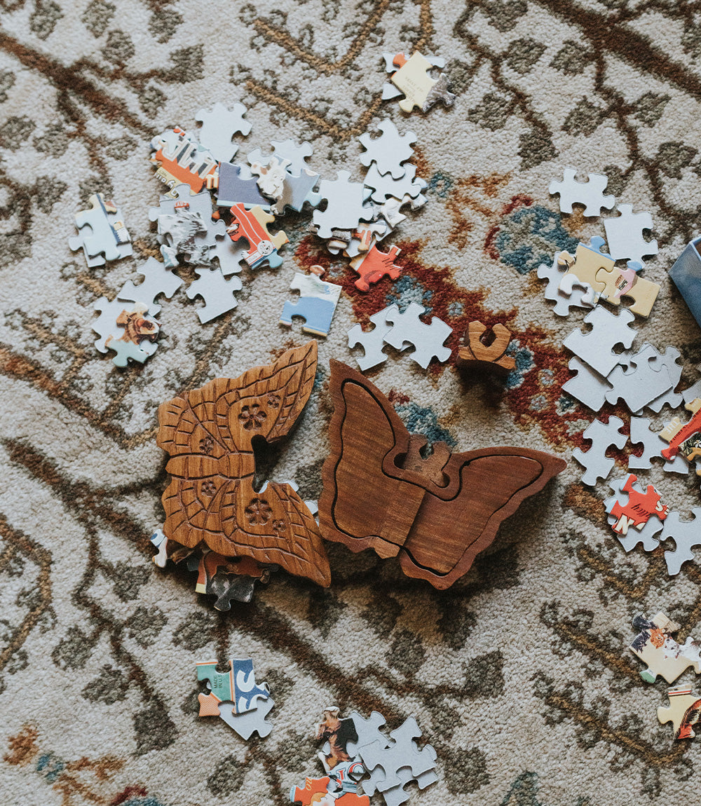 A handcrafted wooden butterfly puzzle box made from solid Indian rosewood with intricate details resembling lace patterns on a carpet surrounded by other jigsaw pieces.