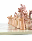 A hand-carved soapstone chess set with a green and pink checkered board and beige, pink, and white stone pieces zoomed in on the white pieces zoomed in on the pink pieces.