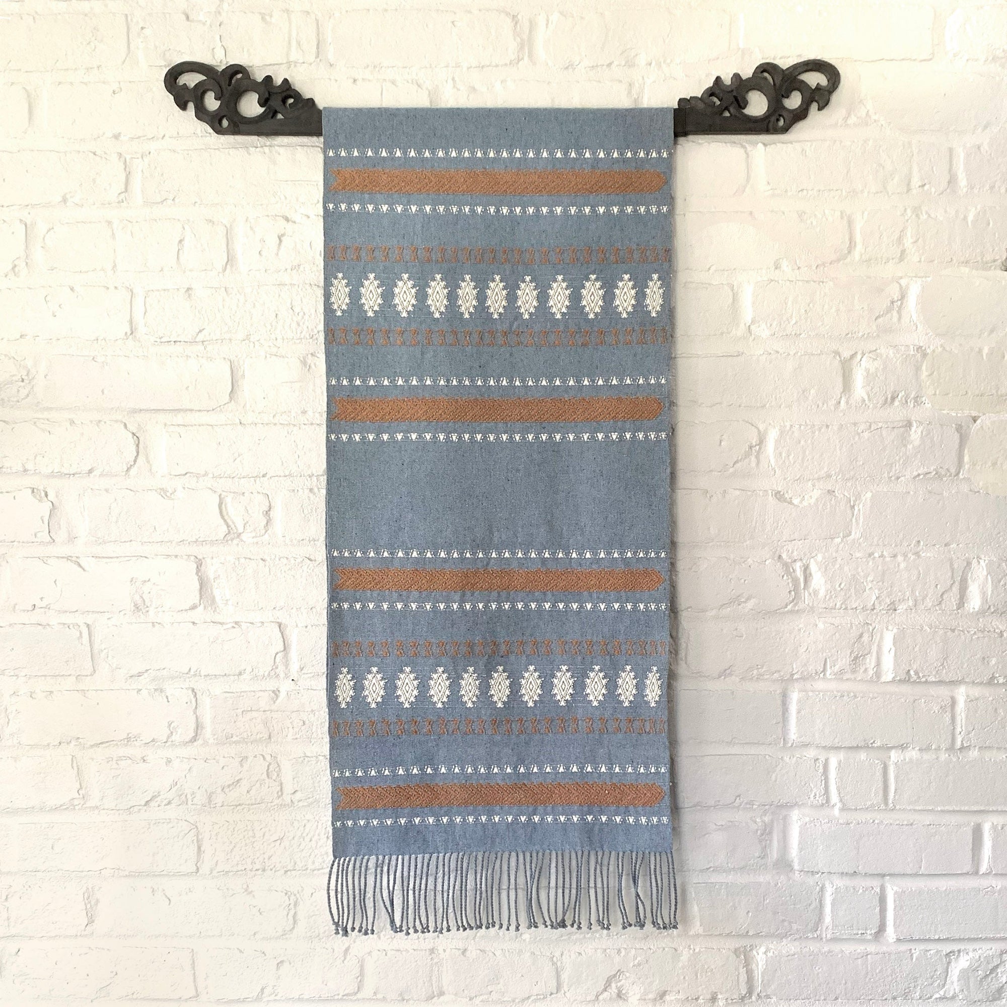 Woven blanket with striped pattern hanging on a decorative black metal hook against a white brick wall.
