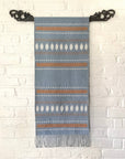 Woven blanket with striped pattern hanging on a decorative black metal hook against a white brick wall.
