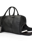 A black leather weekender bag with a leather handle and visible stitching details. Shoes are popping out of a side compartment.