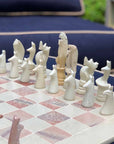 A hand-carved soapstone chess set with a green and pink checkered board and beige, pink, and white stone pieces zoomed in on the white pieces on a couch.
