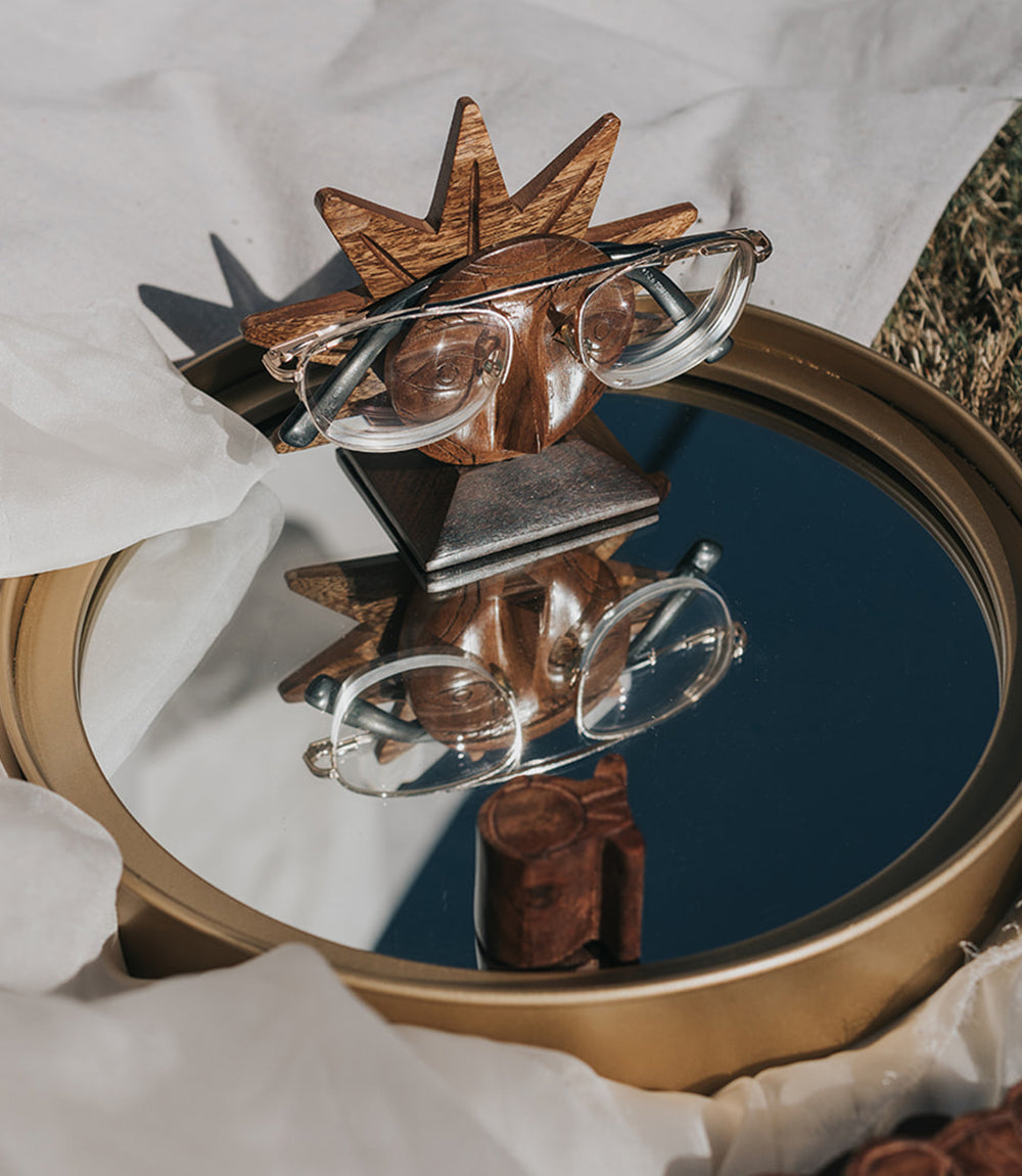 A wooden glasses holder with a sun face design, holding a pair of eyeglasses on a glass tray