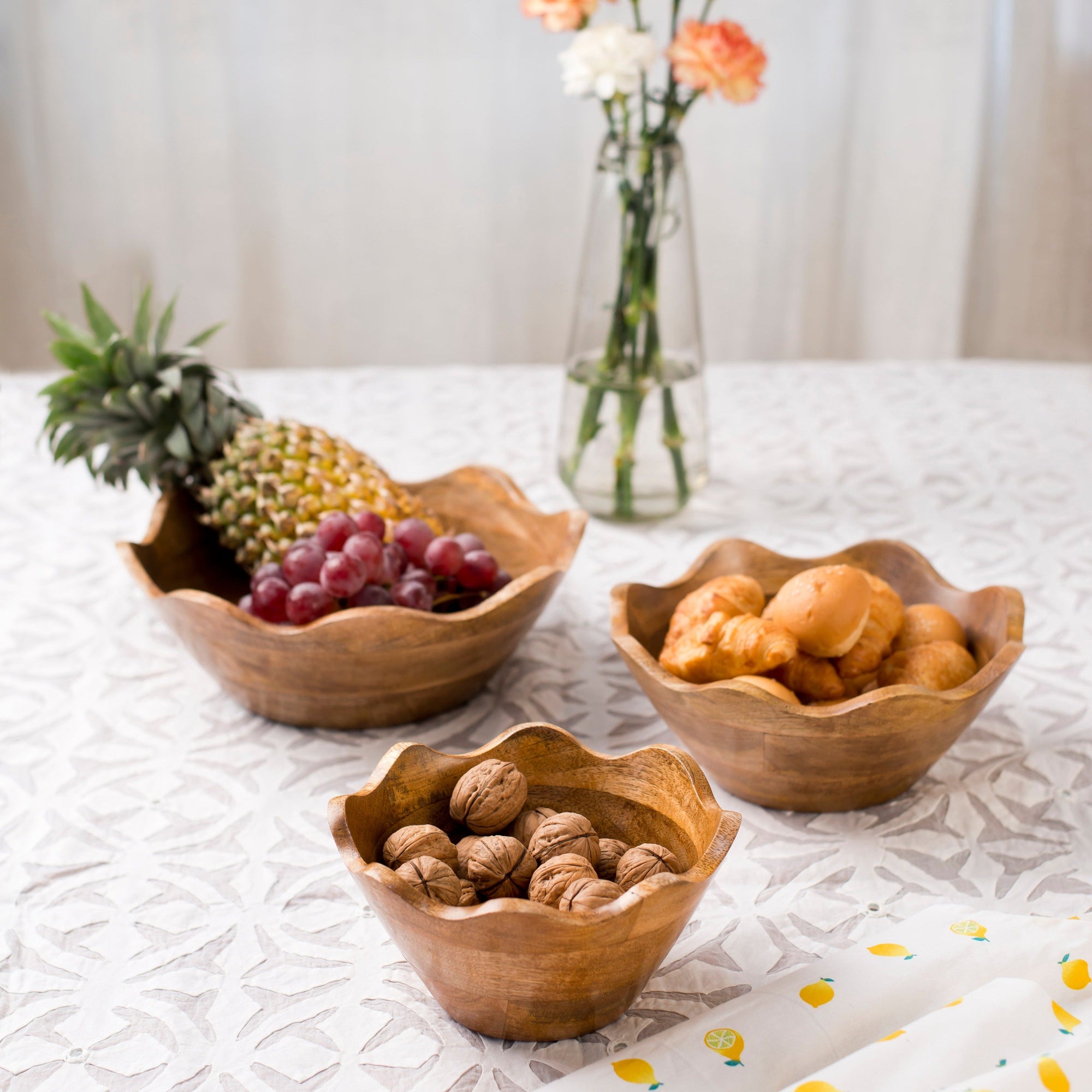 Three wooden bowls containing fruits and nuts on a table with a vase of flowers in the background