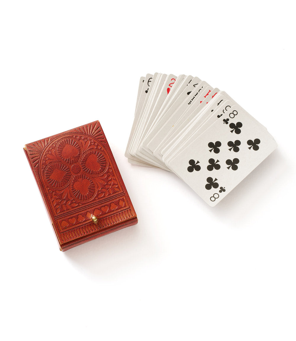 A closed embossed leather box in brown color with intricate floral patterns on the cover alongside the playing cards that go inside of it displayed on a white background.