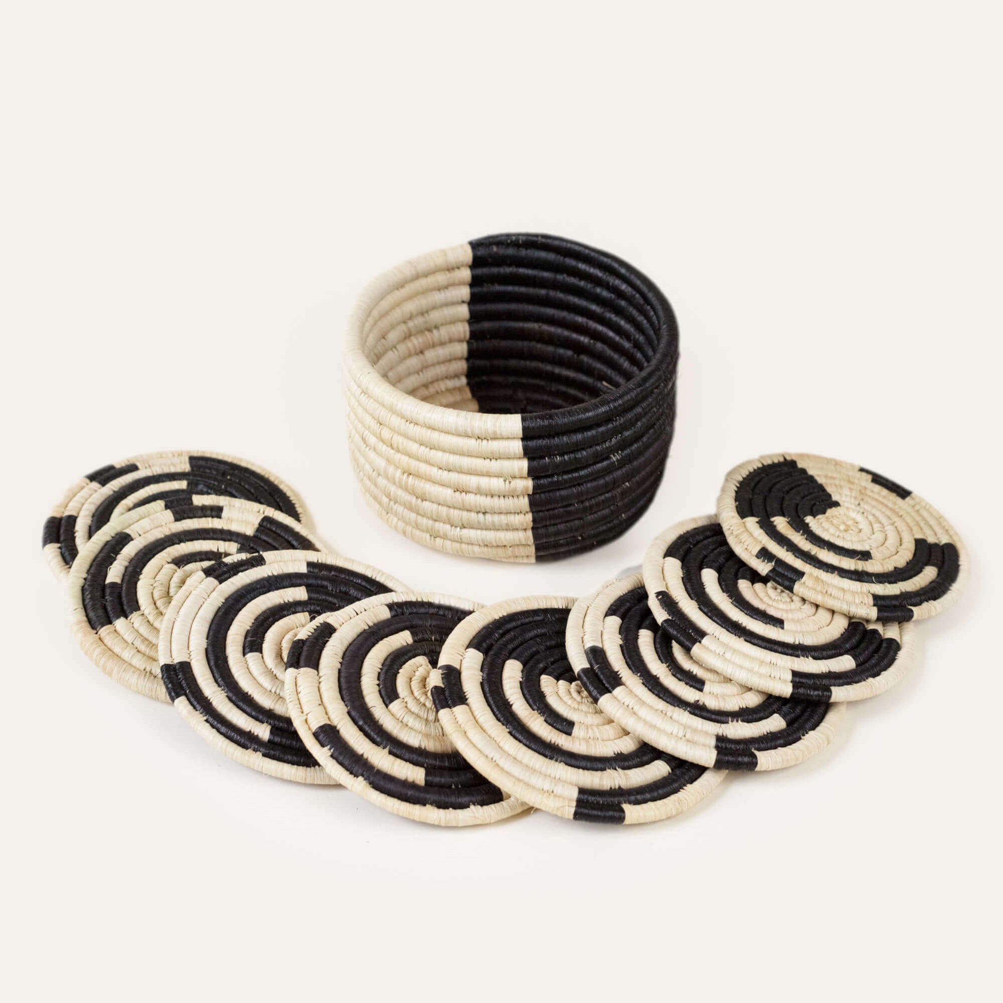 A set of 8 black and natural colored mini basket coasters, displayed in a spiral pattern with one coaster slightly lifted, revealing the others beneath. A small trinket basket is visible in the background