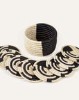 A set of 8 black and natural colored mini basket coasters, displayed in a spiral pattern with one coaster slightly lifted, revealing the others beneath. A small trinket basket is visible in the background