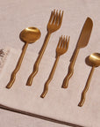Five gold brass flatware pieces, including a dinner knife, dinner fork, soup spoon, and teaspoon on a table with a napkin