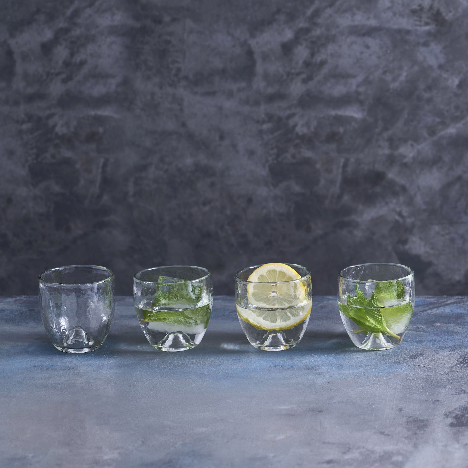 A set of four short, rounded glass tumblers with a distinctive design, displayed in a row against a textured dark background. The glasses appear to be made from partially recycled glass and have a punt in the base as a design element.