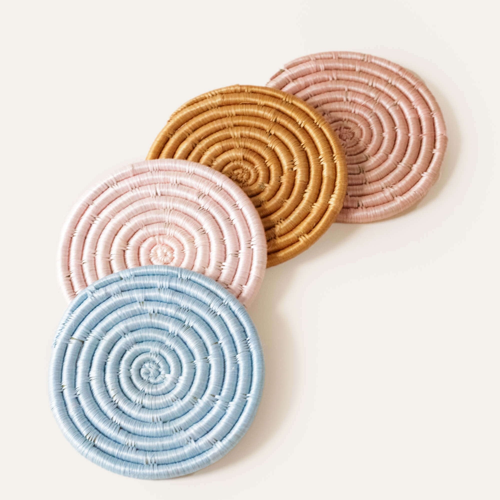 A set of four handcrafted coasters in blue, pink, mauve, and tan colors, made from strands of sisal wrapped around bunches of sweetgrass