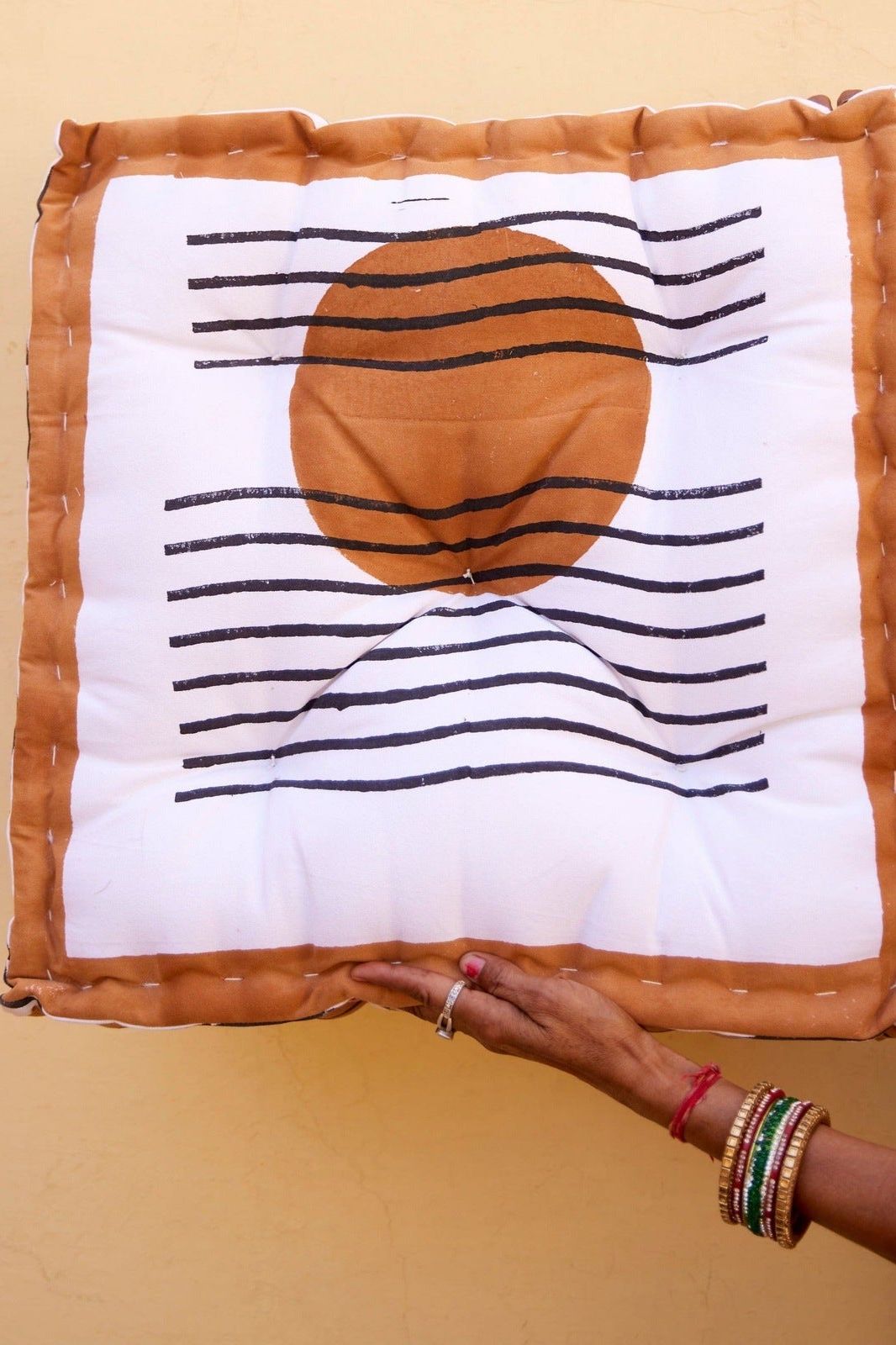 A hand holding a square-shaped floor cushion with a geometric design, predominantly in shades of white, brown, and black