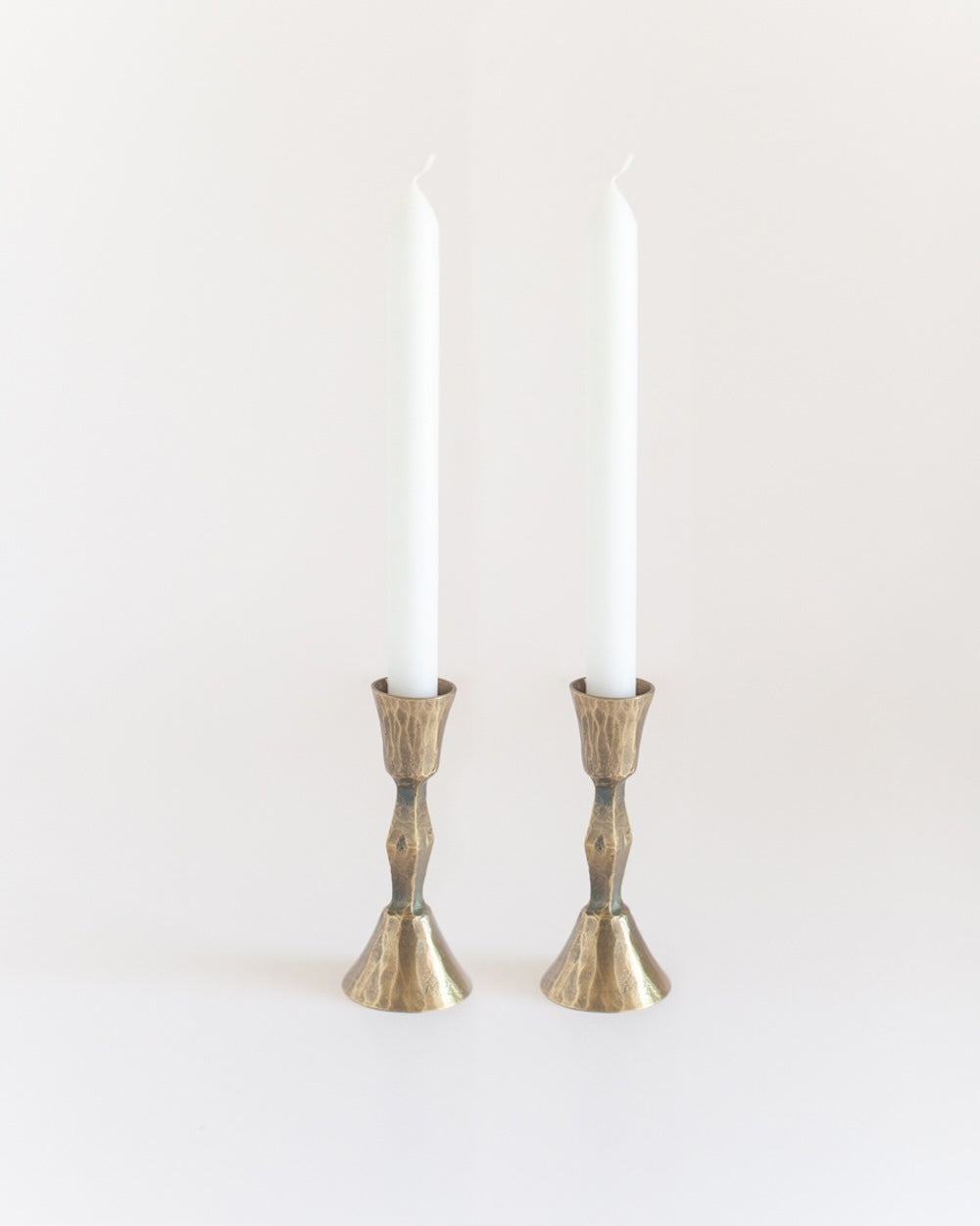 A pair of hand-forged iron candle holders with a brass finish holding candles and featuring a textured design.