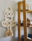 Decorative cotton wall piece with braided design on a wall next to a wooden shelf.