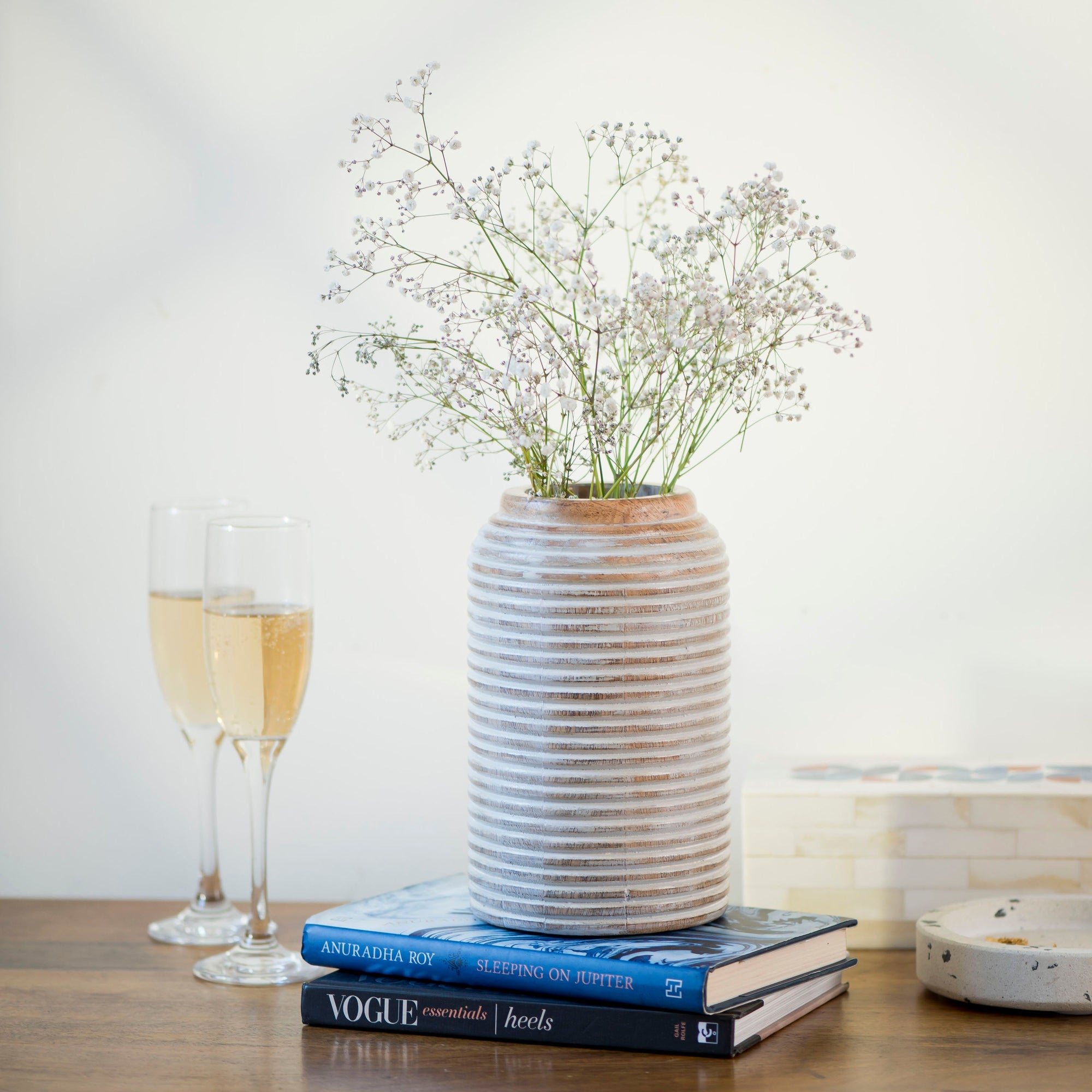 A wooden cylindrical rustic vase with a carved lines design holding flowers on top of two books on top of a table