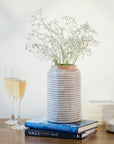 A wooden cylindrical rustic vase with a carved lines design holding flowers on top of two books on top of a table