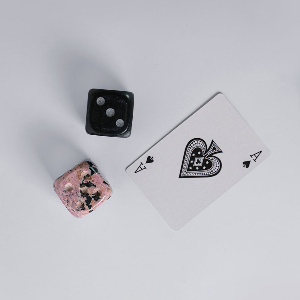 A pair of two hand-carved gemstone dice, in black and pink, displayed on a white surface next to the two of hearts playing card.