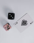 A pair of two hand-carved gemstone dice, in black and pink, displayed on a white surface next to the two of hearts playing card.