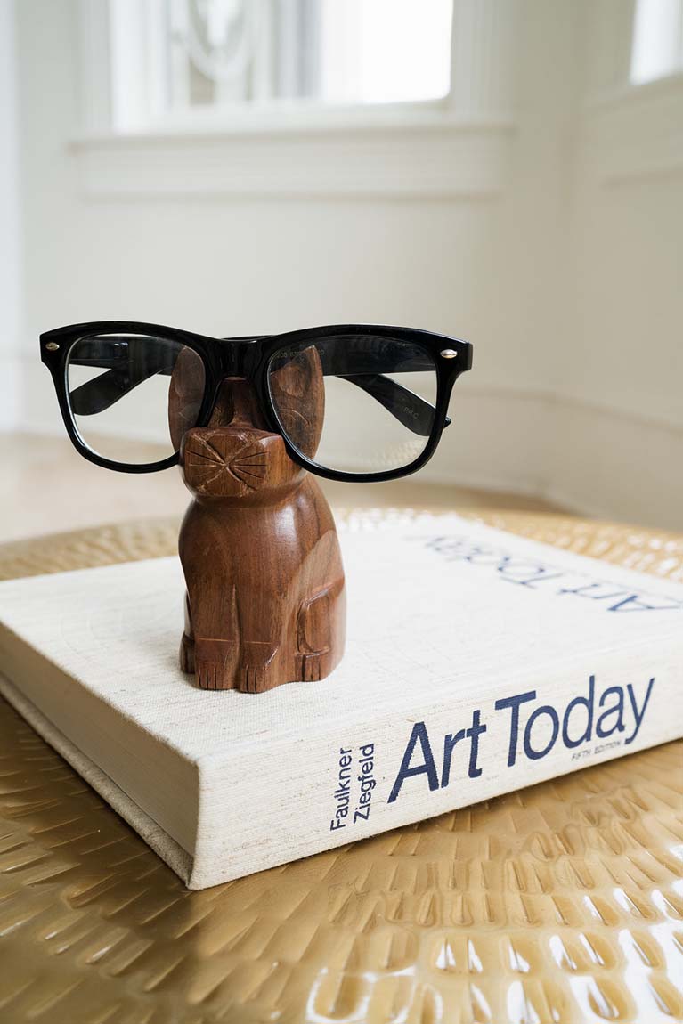 A handmade wooden glasses holder stand in the shape of a cat with eyeglasses on it on top of a book