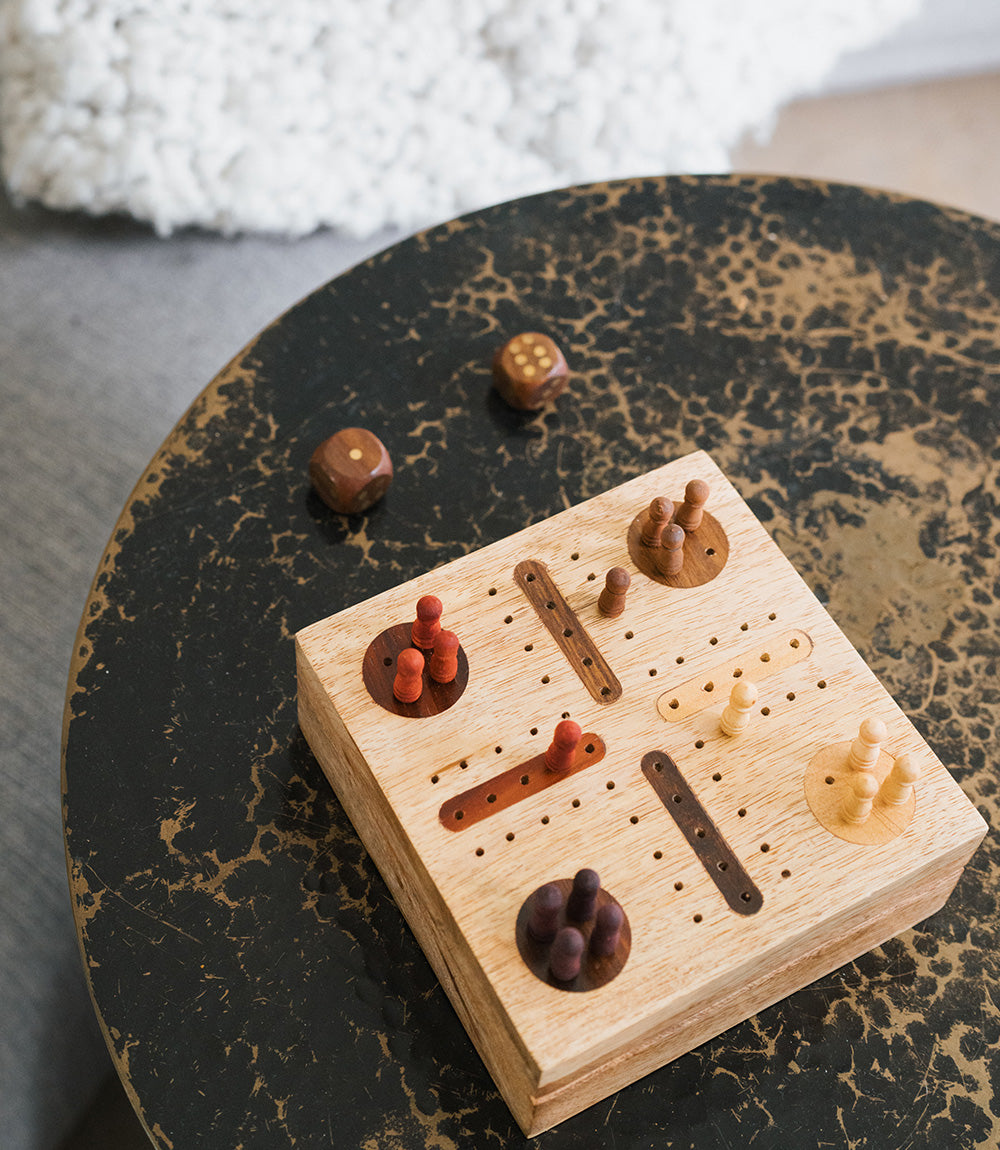 A hand-carved wooden Pachisi game set with a hinged lid, game peg pieces, and dice on a wooden stool.